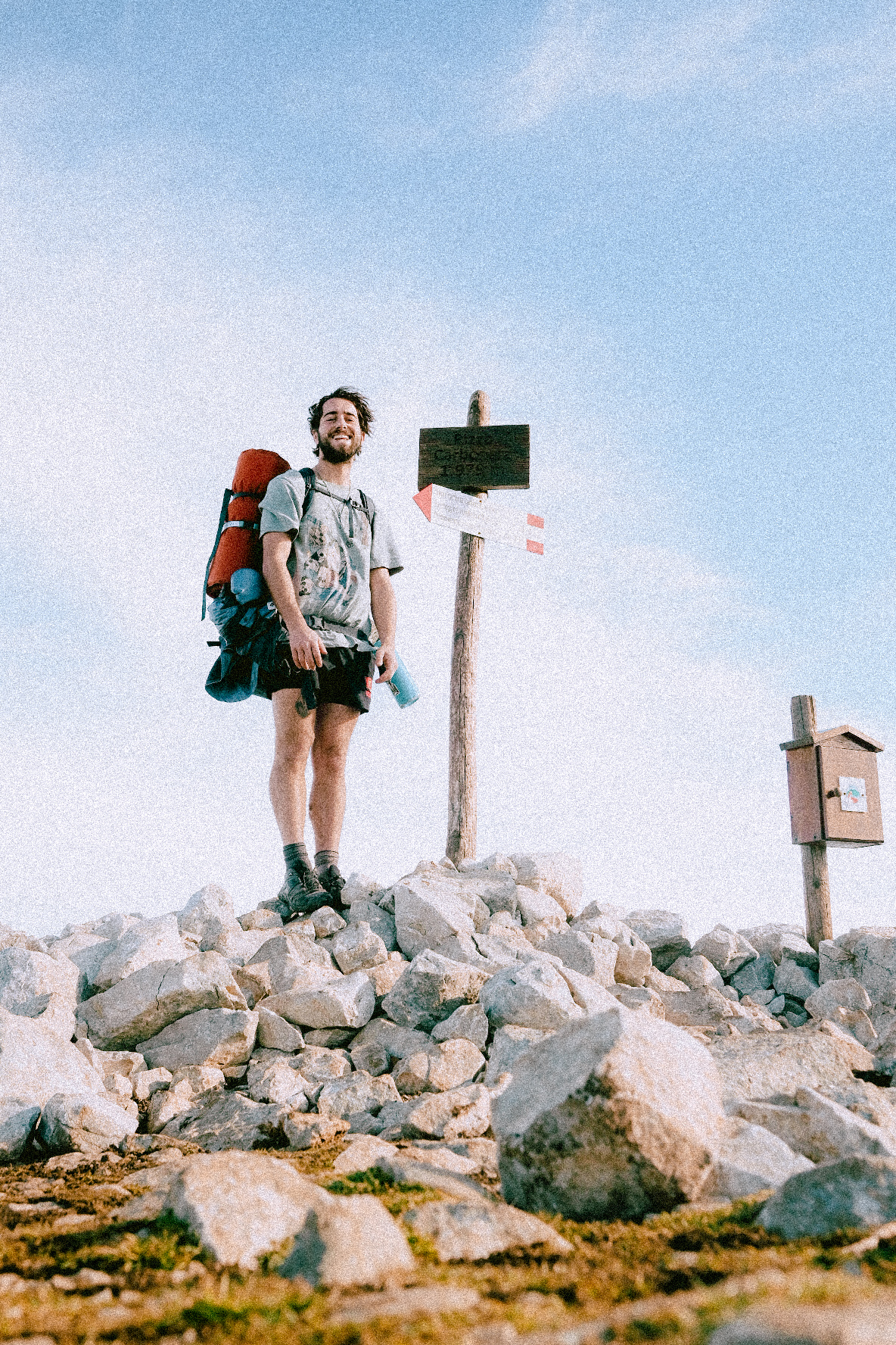 Santo standing at a mountain summit trail marker with a large backpack